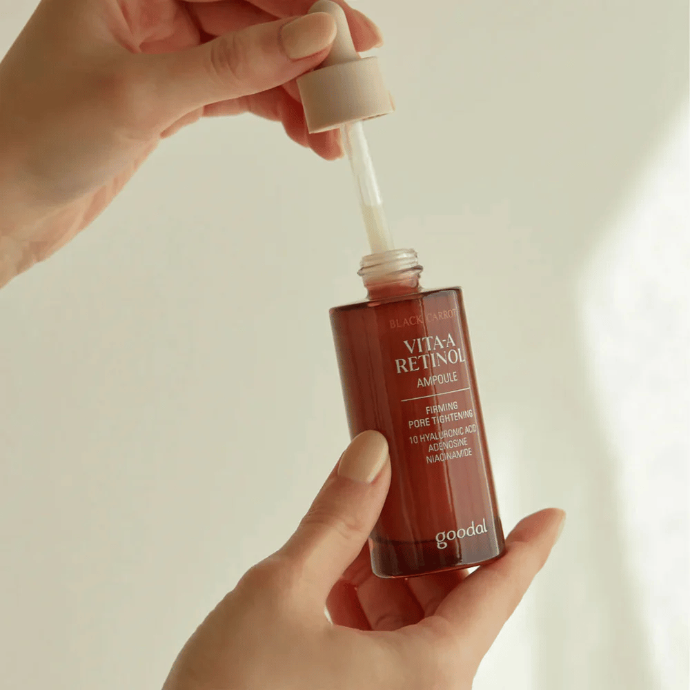 Uma mão segura uma garrafa com Goodal Black Carrot Vita-A Retinol Firming Ampoule. O rótulo da garrafa mostra o nome e as características do produto, incluindo "Firming Pore Tightening" e ingredientes como 10% de ácido hialurônico, adenosina e niacinamida. O conta-gotas é da cor bege.
