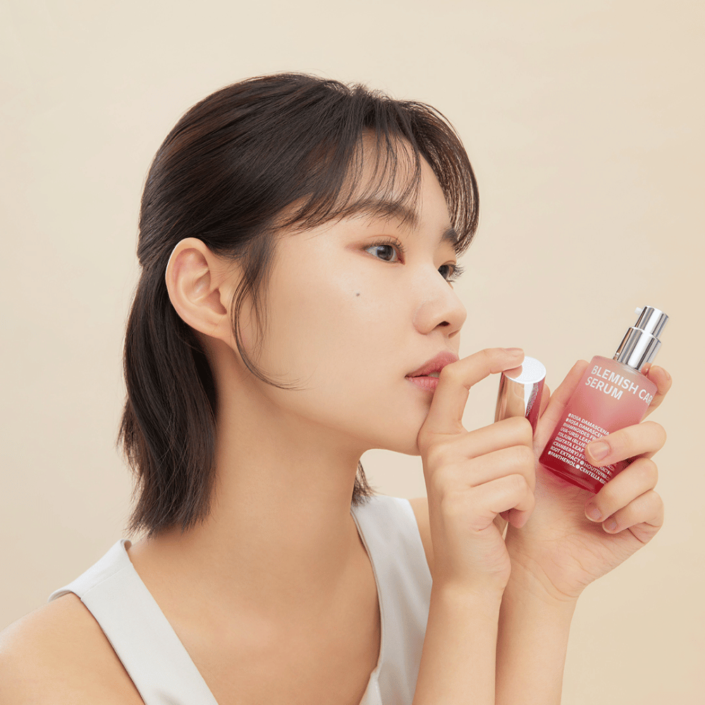 Uma mulher com cabelo escuro segura uma garrafa de Blemish Care Serum em uma mão e tira a tampa com a outra. Ela tem uma expressão facial neutra e o fundo é bege claro, o que dá uma impressão suave e minimalista.