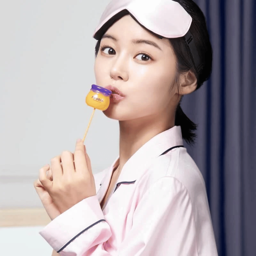 A mulher na imagem está usando um bálsamo labial de mel e mirtilo da FRUDIA, o que sugere uma rotina de cuidados com a pele com ingredientes naturais.