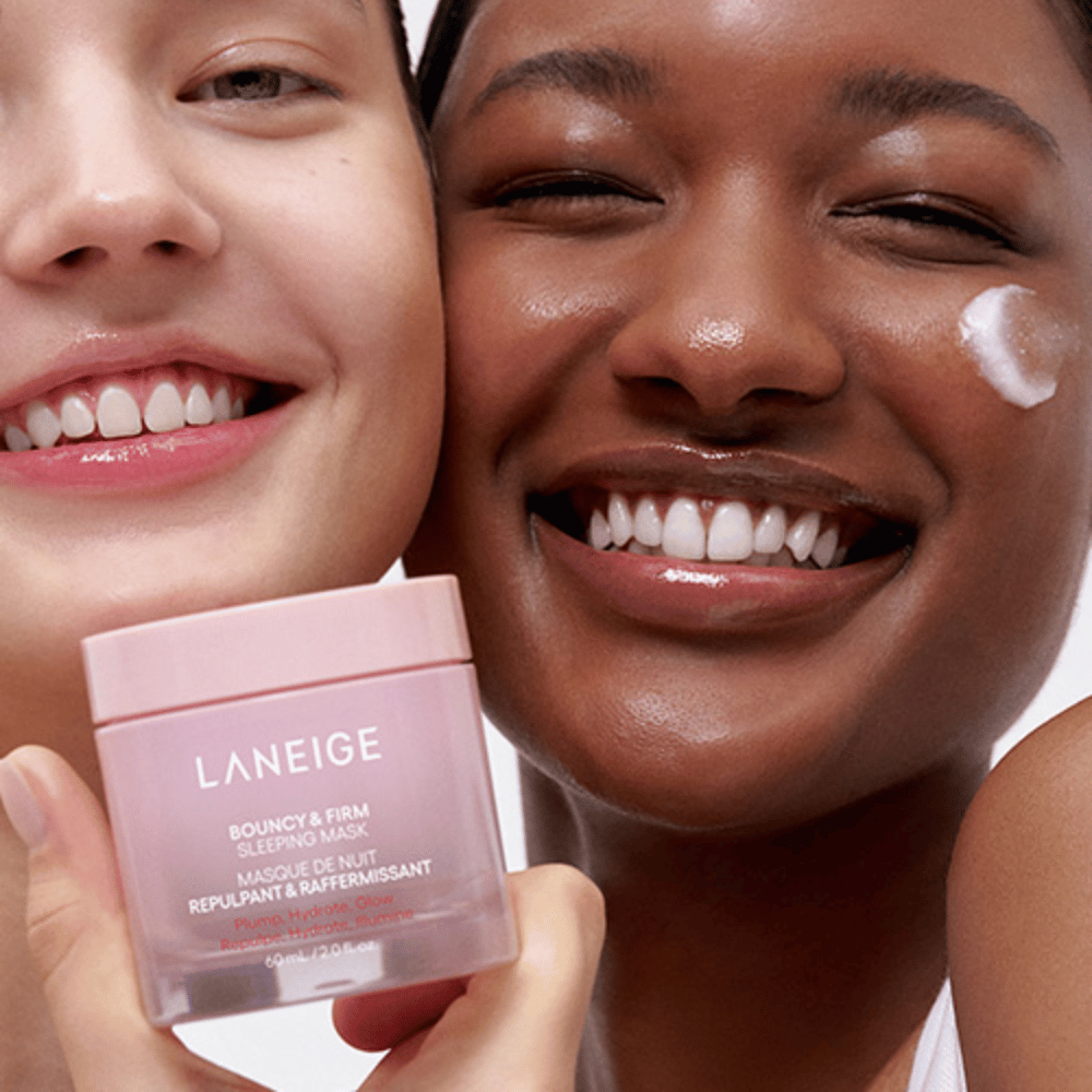 Duas pessoas sorrindo e segurando Laneige Bouncy & Firm Sleeping Mask. Uma delas aplicou a máscara na bochecha, e o produto é claramente visível em suas mãos. A máscara está em um frasco rosa e é comercializada como "Volume, Hidratar, Brilhar."