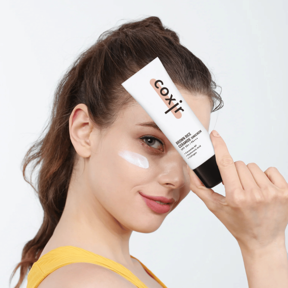 Mulher com protetor solar aplicado na bochecha segura um tubo Coxir Brown Rice Ceramide Sunscreen, SPF 50+ PA+++, contra a testa e sorri levemente.