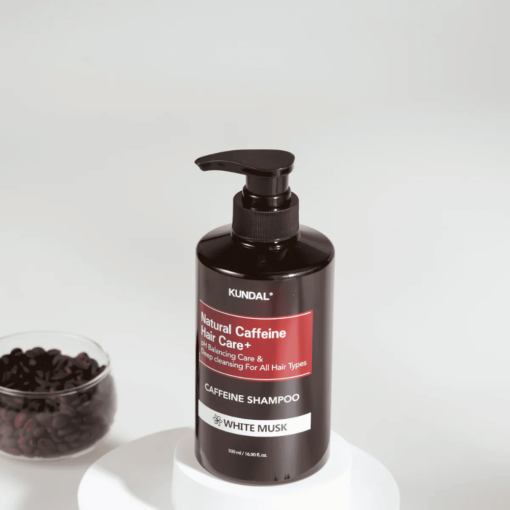 KUNDAL Natural Caffeine Hair Care+ Shampoo White Musk em garrafa preta com bomba, 500 ml, colocado ao lado de uma tigela com grãos de café.