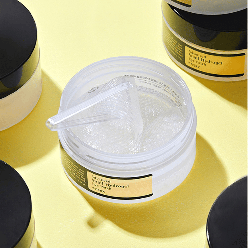 Um frasco aberto COSRX Advanced Snail Hydrogel Eye Patch com adesivos para olhos transparentes e uma pequena espátula, cercado por mais frascos fechados sobre fundo amarelo.
