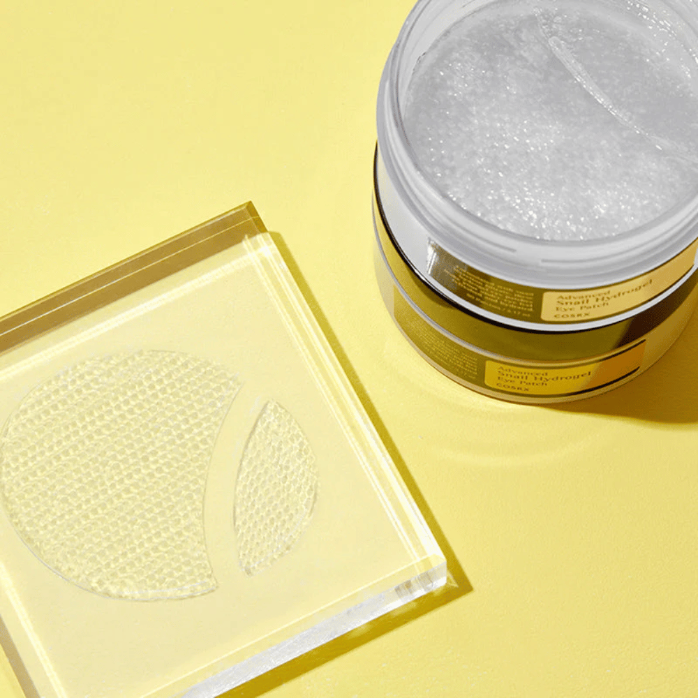 Um frasco aberto COSRX Advanced Snail Hydrogel Eye Patch está sobre um fundo amarelo ao lado de um suporte transparente onde dois adesivos para olhos estão colocados.