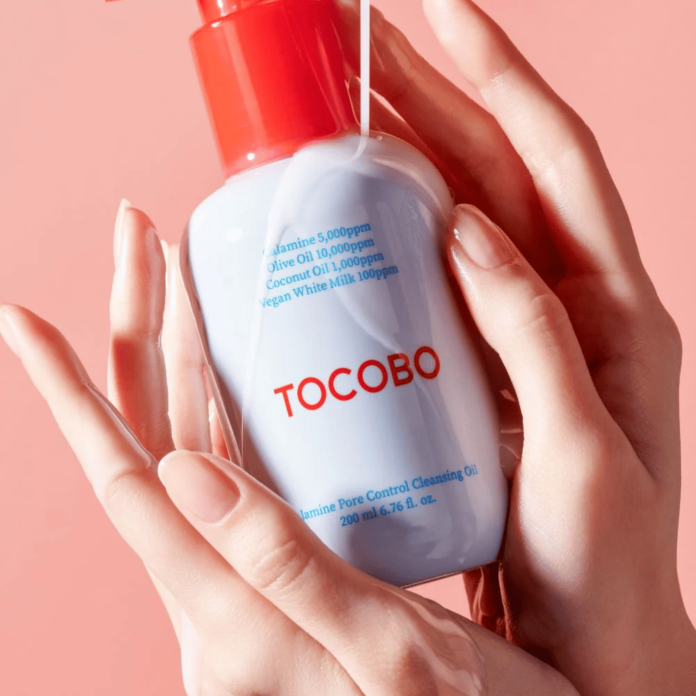 TOCOBO Calamine Pore Control Cleansing Oil segurado nas mãos contra um fundo rosa, a bomba vermelha do frasco e a lista de ingredientes são claramente visíveis.