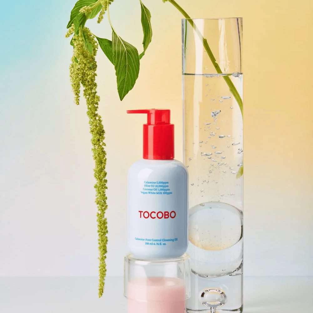 TOCOBO Calamine Pore Control Cleansing Oil com bomba vermelha está ao lado de um tubo de vidro alto cheio de água e uma planta verde, contra um fundo de cor pastel clara.