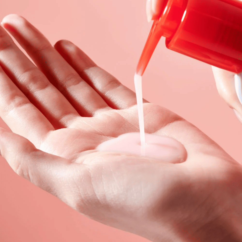 Um óleo de limpeza rosa claro é aplicado de um frasco de bomba vermelho em uma mão aberta contra um fundo rosa.