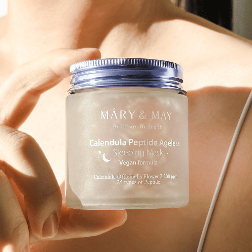 Uma mulher segura um frasco com MARY & MAY Calendula Peptide Ageless Sleeping Mask. O frasco é transparente com uma tampa roxa e o texto "Calendula Peptide Ageless Sleeping Mask" é claramente visível. O produto é uma máscara de dormir vegana com calêndula e peptídeos, destinada a dar à pele um brilho jovem durante a noite.