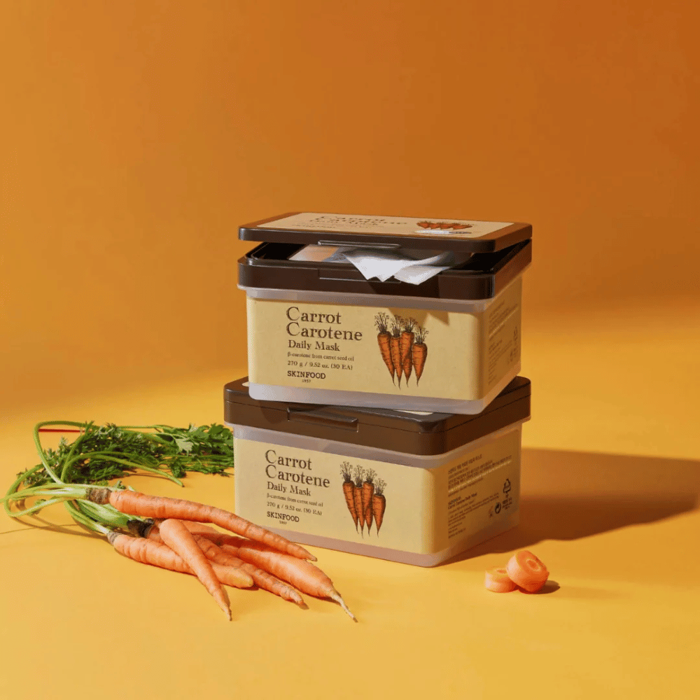 A imagem mostra três embalagens empilhadas de "Carrot Carotene Daily Mask" de SKINFOOD, colocadas sobre um fundo laranja junto com cenouras frescas. Cada caixa contém máscaras faciais enriquecidas com β-caroteno de óleo de semente de cenoura, projetadas para uso diário para nutrir e cuidar da pele.