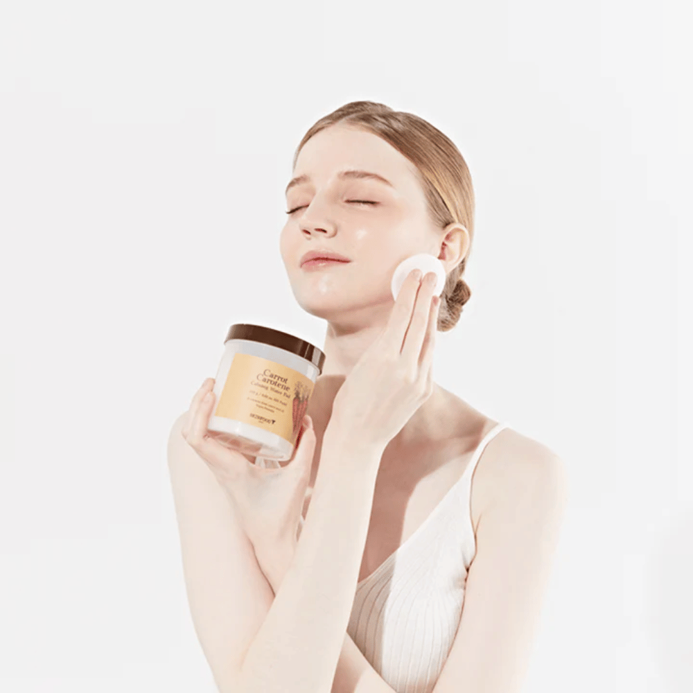 Uma mulher com cabelo preso aplica um produto de cuidados com a pele no rosto com um disco de algodão. Ela segura um grande frasco marcado "Carrot Carotene Calming Water Pad". Seus olhos fechados e expressão pacífica transmitem uma sensação de relaxamento e prazer na rotina de cuidados com a pele.