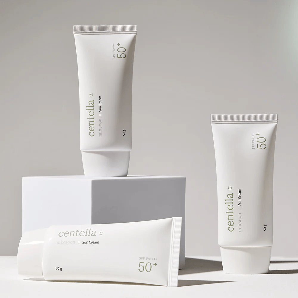 Três embalagens de Centella Sun Cream SPF 50+ PA++++ organizadas esteticamente em pódios brancos. Dois tubos estão em pé e um está deitado, todos com design branco minimalista e detalhes de texto verdes.