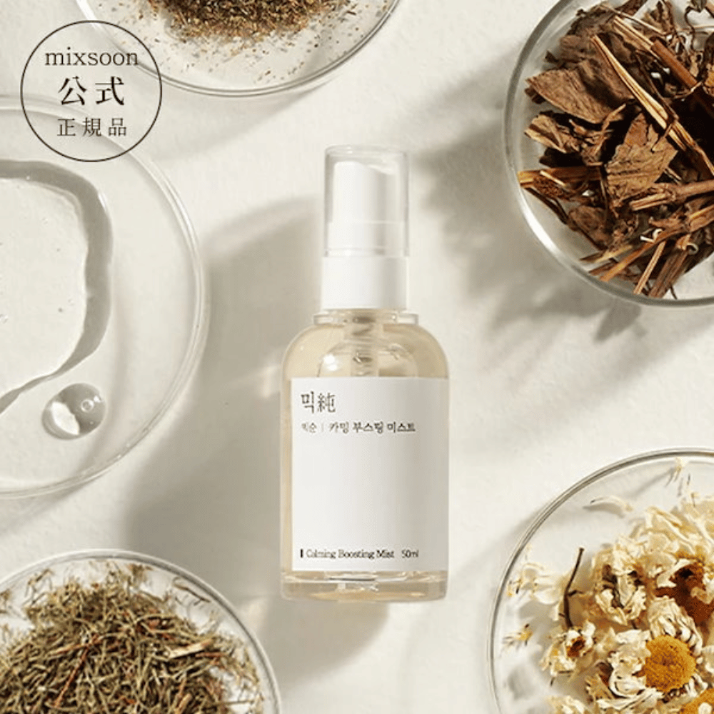 Um frasco de spray transparente com o rótulo 'Calming Boosting Mist' da Mixsoon, rodeado por ervas secas e flores em tigelas brancas contra um fundo bege, destacando os ingredientes naturais dos produtos.
