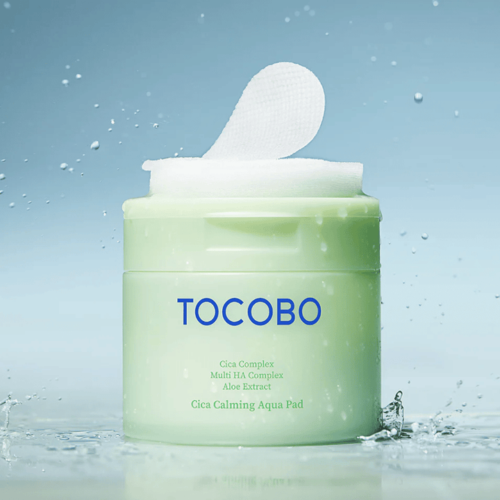 Frasco aberto com TOCOBO Cica Calming Aqua Pad, onde um disco tónico sobressai da embalagem. Gotas de água à volta do produto reforçam a sensação de hidratação intensa e frescura.