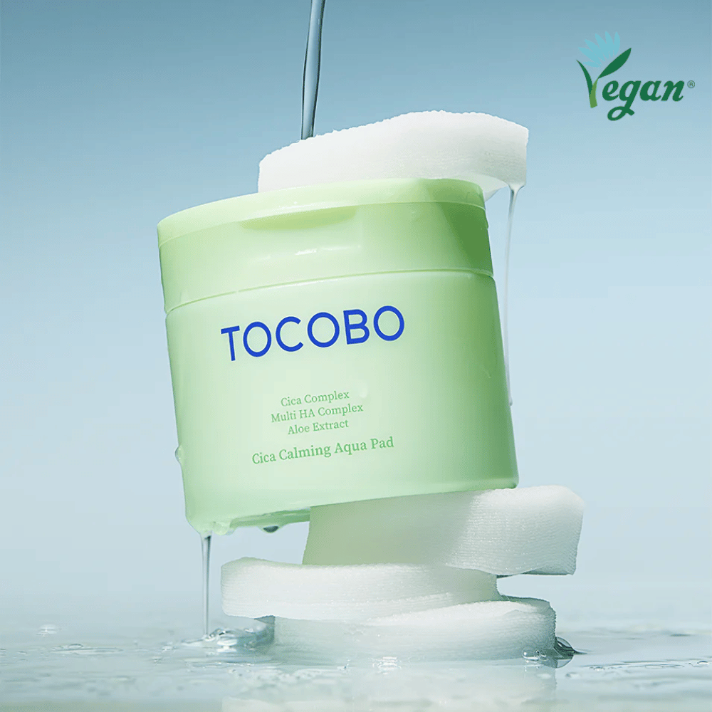 TOCOBO Cica Calming Aqua Pad colocado entre discos tónicos com essência a escorrer. O símbolo vegan destaca a formulação vegana do produto e o seu perfil amigo da pele.