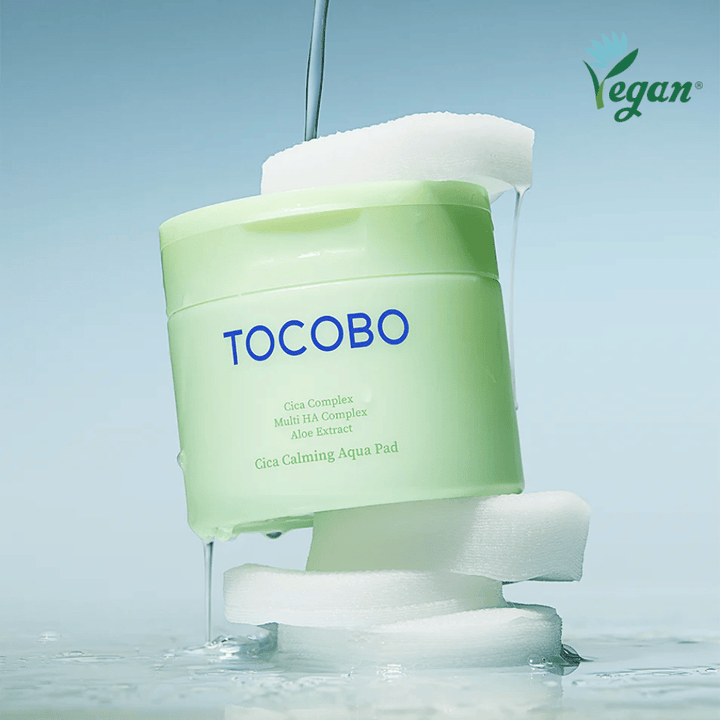 TOCOBO Cica Calming Aqua Pad colocado entre discos tónicos com essência a escorrer. O símbolo vegan destaca a formulação vegana do produto e o seu perfil amigo da pele.