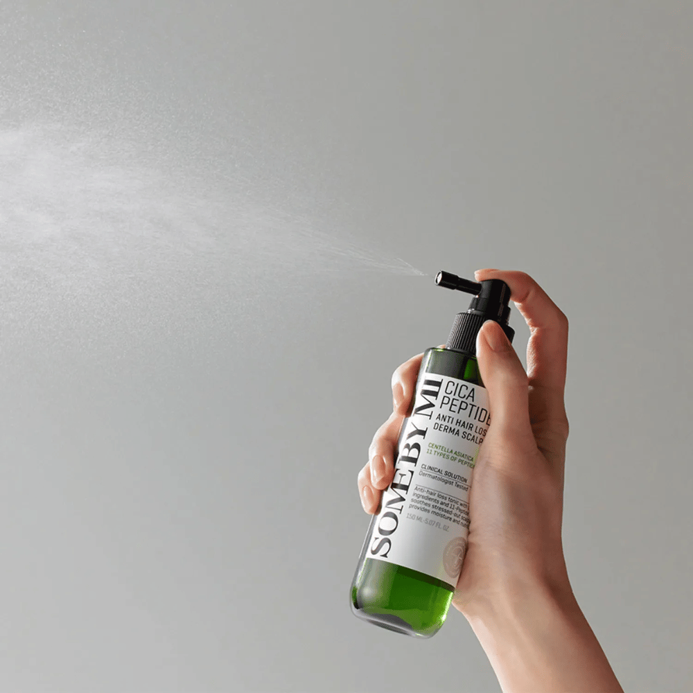 Mão pulverizando SOME BY MI Cica Peptide Anti Hair Loss Derma Scalp Tonic de uma garrafa verde com bico preto e rótulo branco contra fundo cinza.