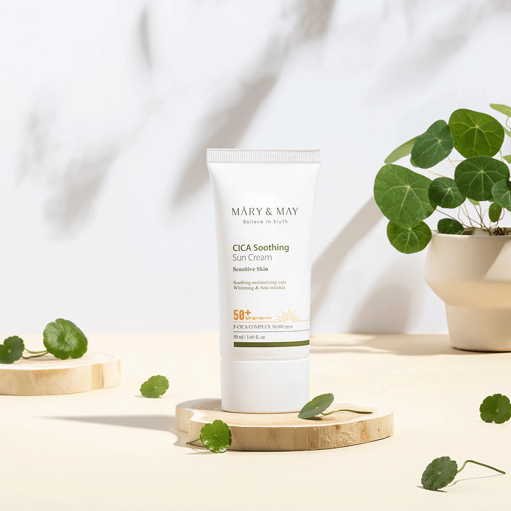 Mary&May CICA Soothing Sun Cream colocada sobre um pódio de madeira com folhas verdes de centella à sua volta. A imagem enfatiza os ingredientes calmantes para a pele e a inspiração natural por detrás do produto.