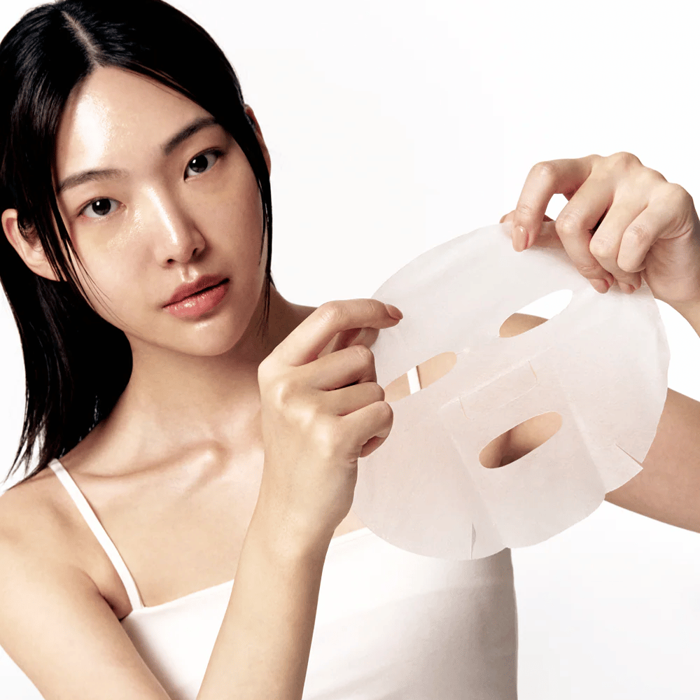 Mulher com pele limpa e hidratada segura uma Sheet mask branca à sua frente, pronta para aplicá-la no rosto.