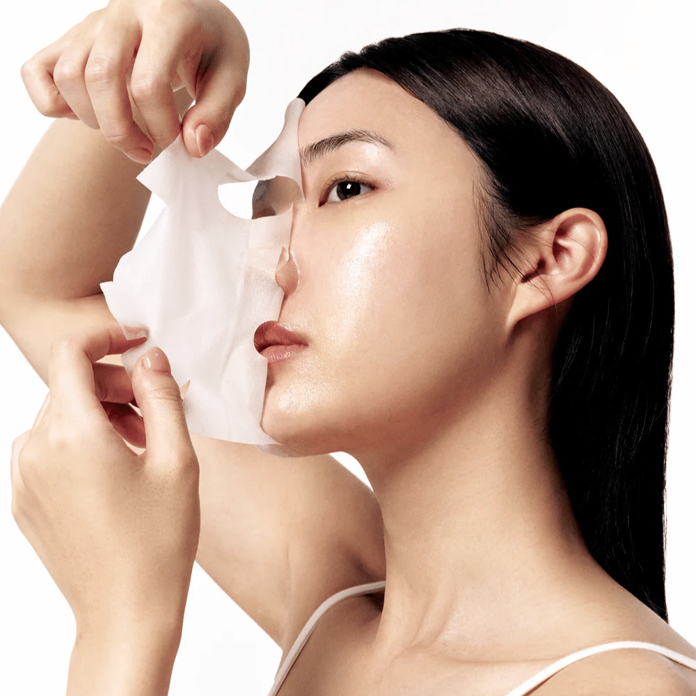 Mulher com pele lisa e hidratada remove uma Sheet mask branca do rosto, revelando um tom de pele radiante e uniforme.