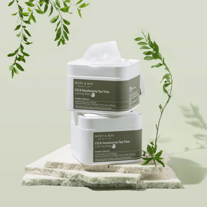 Två burkar Mary & May Cica Houttuynia Tea Tree Calming Mask placerade på natursten med gröna växtdetaljer – lugnande sheetmasker för känslig hud.