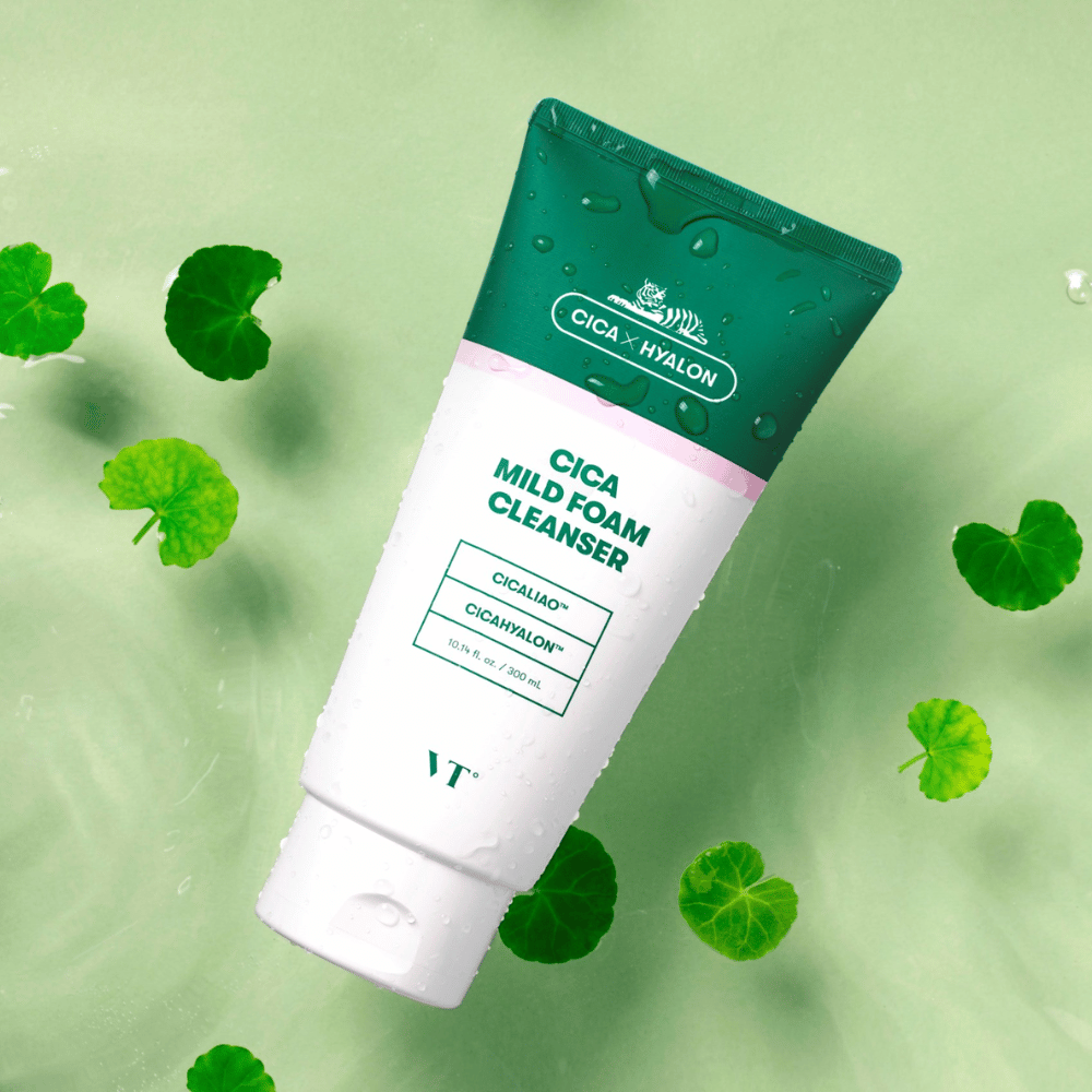 VT Cica Mild Foam Cleanser deitado sobre uma superfície verde clara com gotas de água na embalagem e rodeado por folhas verdes de centella asiatica, destacando as propriedades calmantes do produto.