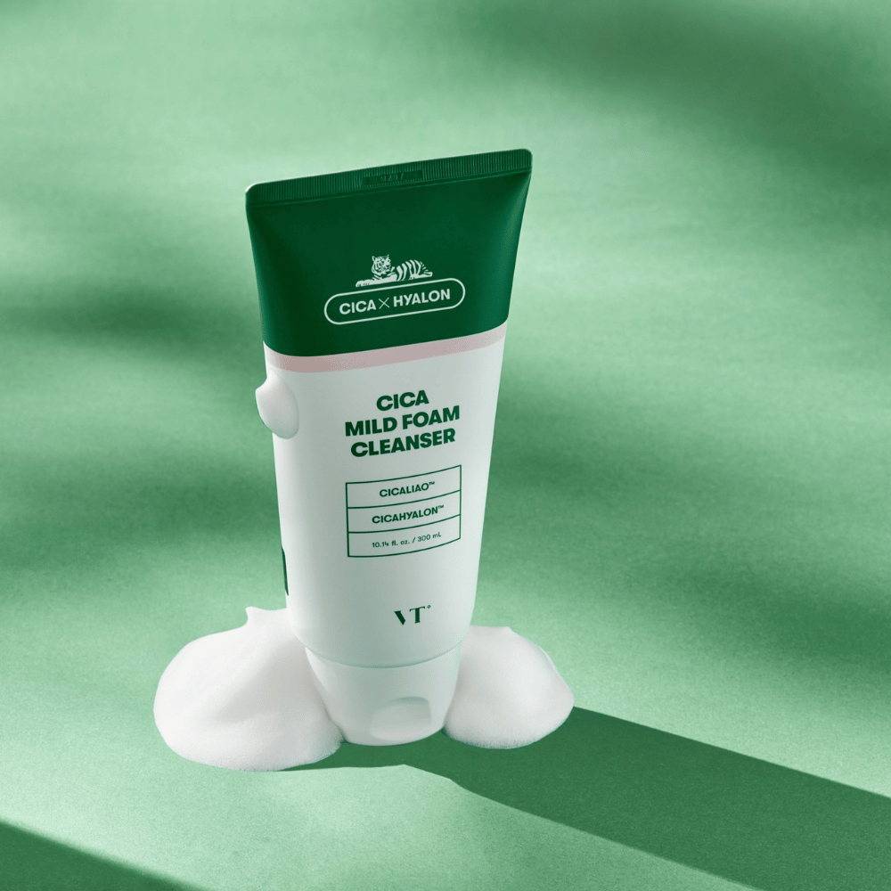 Tubo com VT Cica Mild Foam Cleanser em pé sobre uma superfície verde com espuma ao redor da base. O produto tem design verde e branco e é comercializado com CICALIAO™ e CICAHYALON™.
