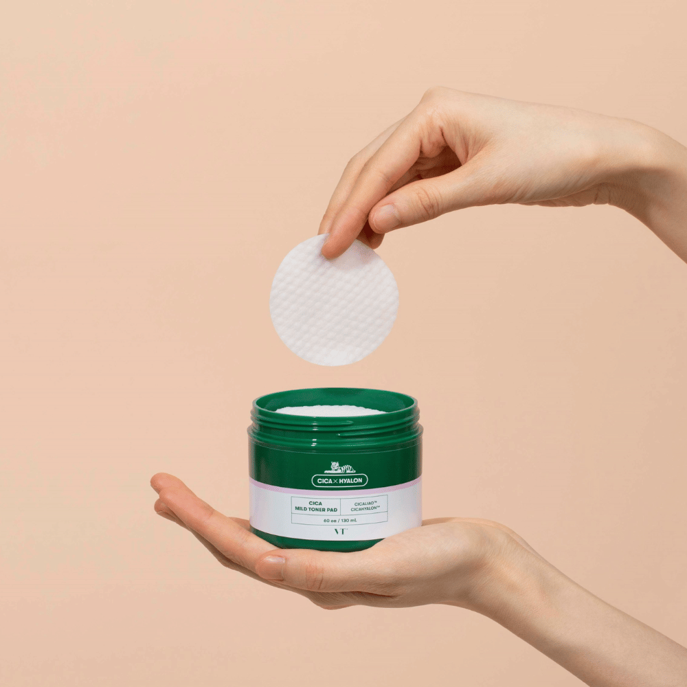 Mão segurando um frasco verde com VT Cica Mild Toner Pad e levantando uma almofada de toner redonda e branca sobre o frasco contra fundo bege.