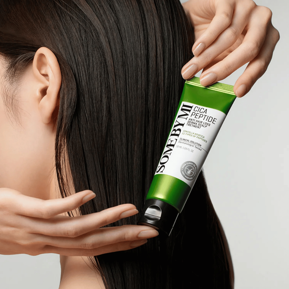 Mulher aplica SOME BY MI Cica Peptide Anti Hair Loss Derma Scalp Treatment em cabelo longo e escuro com um tubo verde e branco.