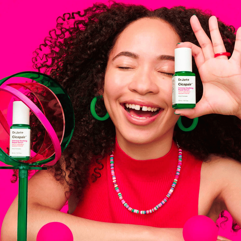 Uma mulher sorri amplamente enquanto segura Dr.Jart+ Cicapair Intensive Soothing Repair Serum em frente ao seu rosto. Ela usa brincos verdes e um colar vermelho. O fundo é rosa claro e decorado com círculos verdes e rosa.