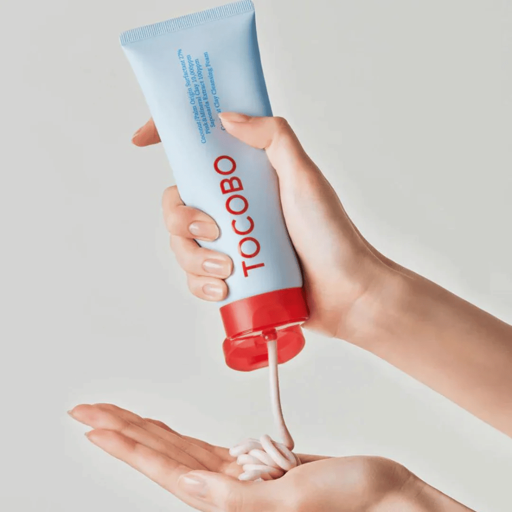Uma pessoa segura um tubo de TOCOBO Coconut Clay Cleansing Foam. O tubo é azul claro com uma tampa vermelha e o texto "TOCOBO" na frente. O produto é espremido do tubo e moldado como um shampoo na mão. O fundo é neutro e dá foco ao produto.
