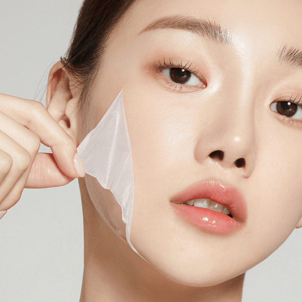 A imagem mostra uma jovem removendo uma máscara facial fina, destacando sua pele hidratada e macia. A máscara segue os contornos do rosto, dando a impressão de um produto de cuidados com a pele eficaz e suave. Sua expressão é relaxada.