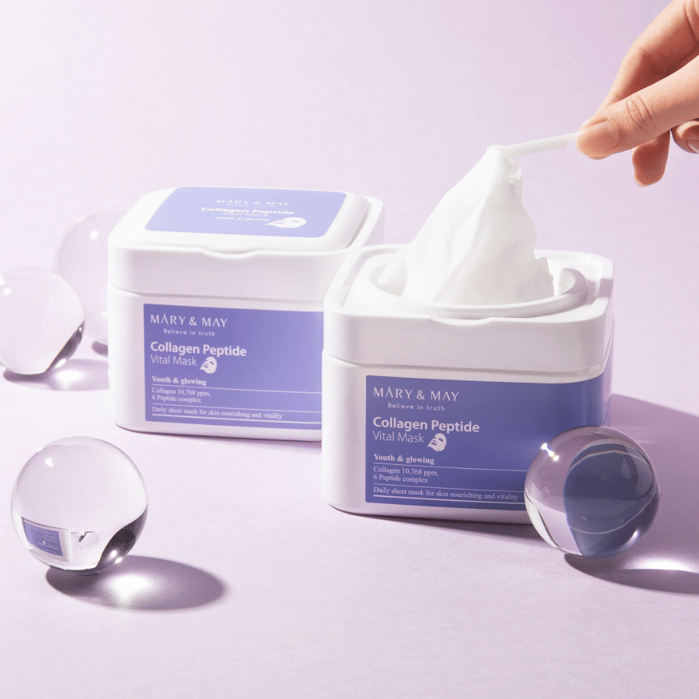 Uma mão pega uma máscara de um frasco da MARY & MAY Collagen Peptide Vital Mask. A máscara é destinada ao uso diário para nutrir e dar brilho à pele, com colágeno e peptídeos para apoiar a juventude da pele.