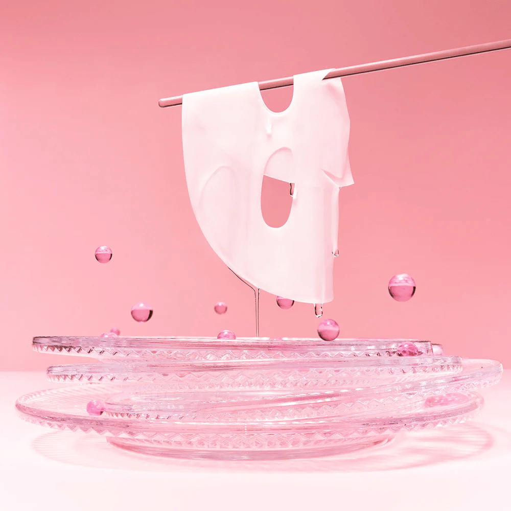Uma máscara de hidrogel está pendurada em uma haste, pingando com soro. O fundo é rosa e o espelho na parte inferior reflete as bolhas rosas que cercam a máscara.