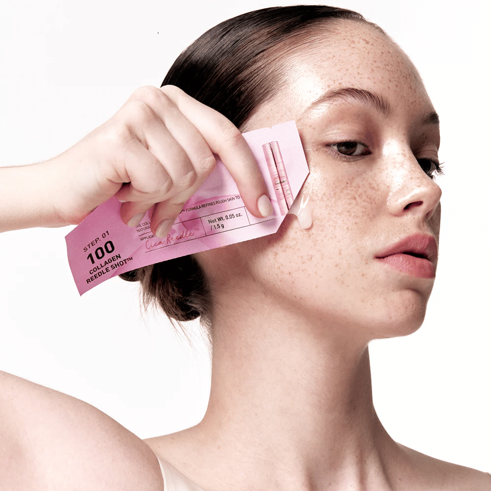 Mulher aplica o passo 1 de Collagen Reedle Shot 100 com uma embalagem rosa descartável na bochecha. Um gel claro é visível na pele, marcando o primeiro passo do tratamento de cuidados com a pele em duas etapas.