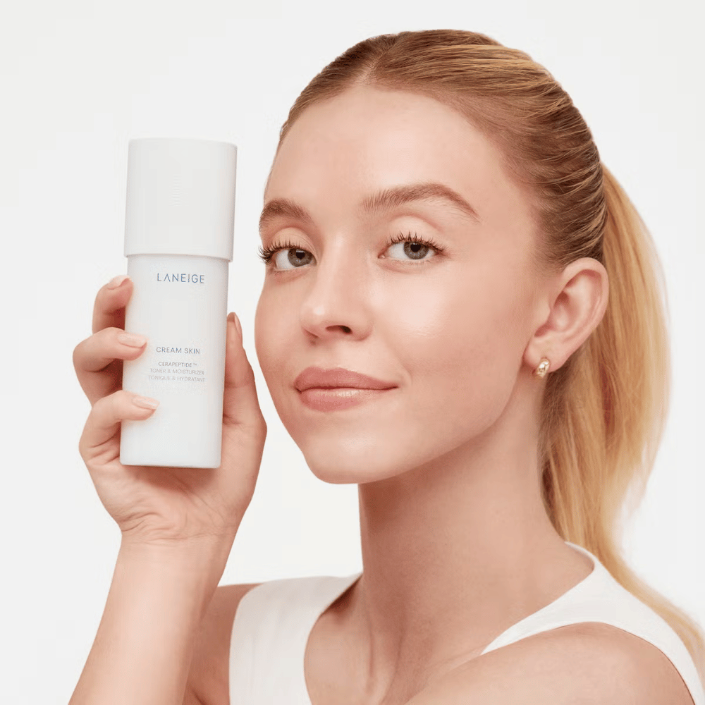 Uma pessoa segura Laneige Cream Skin Cerapeptide Refiner em uma garrafa branca. A pessoa sorri e mostra o produto, que é comercializado como um tônico hidratante e produto de cuidado da pele.