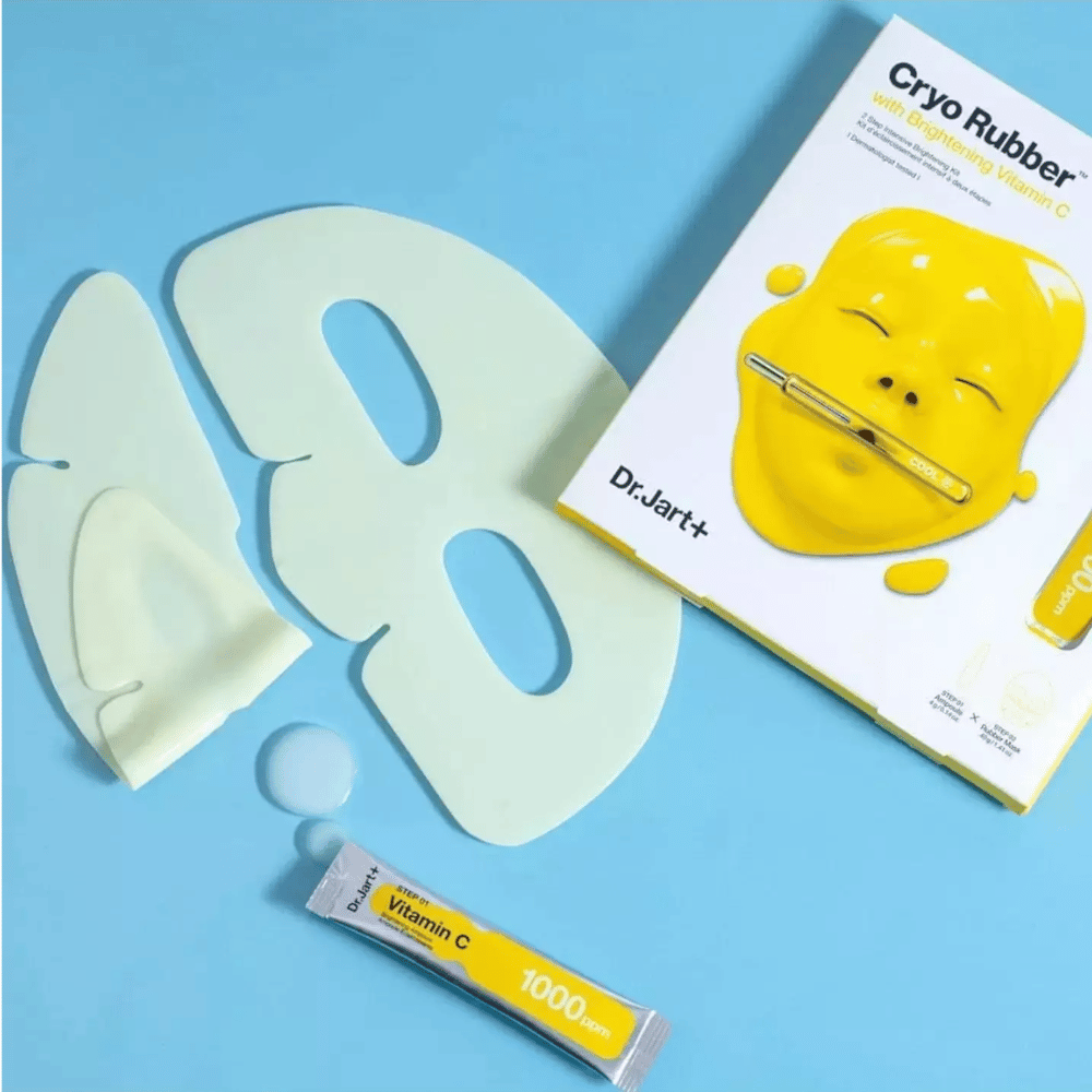 Imagem do produto de beleza 'Cryo Rubber™ com Vitamina C Iluminadora' de Dr.Jart+. Em um fundo azul, vê-se uma máscara facial de borracha branca, um pequeno tubo marcado 'Vitamina C 1000ppm', e uma caixa com o nome do produto e imagem da máscara amarela.