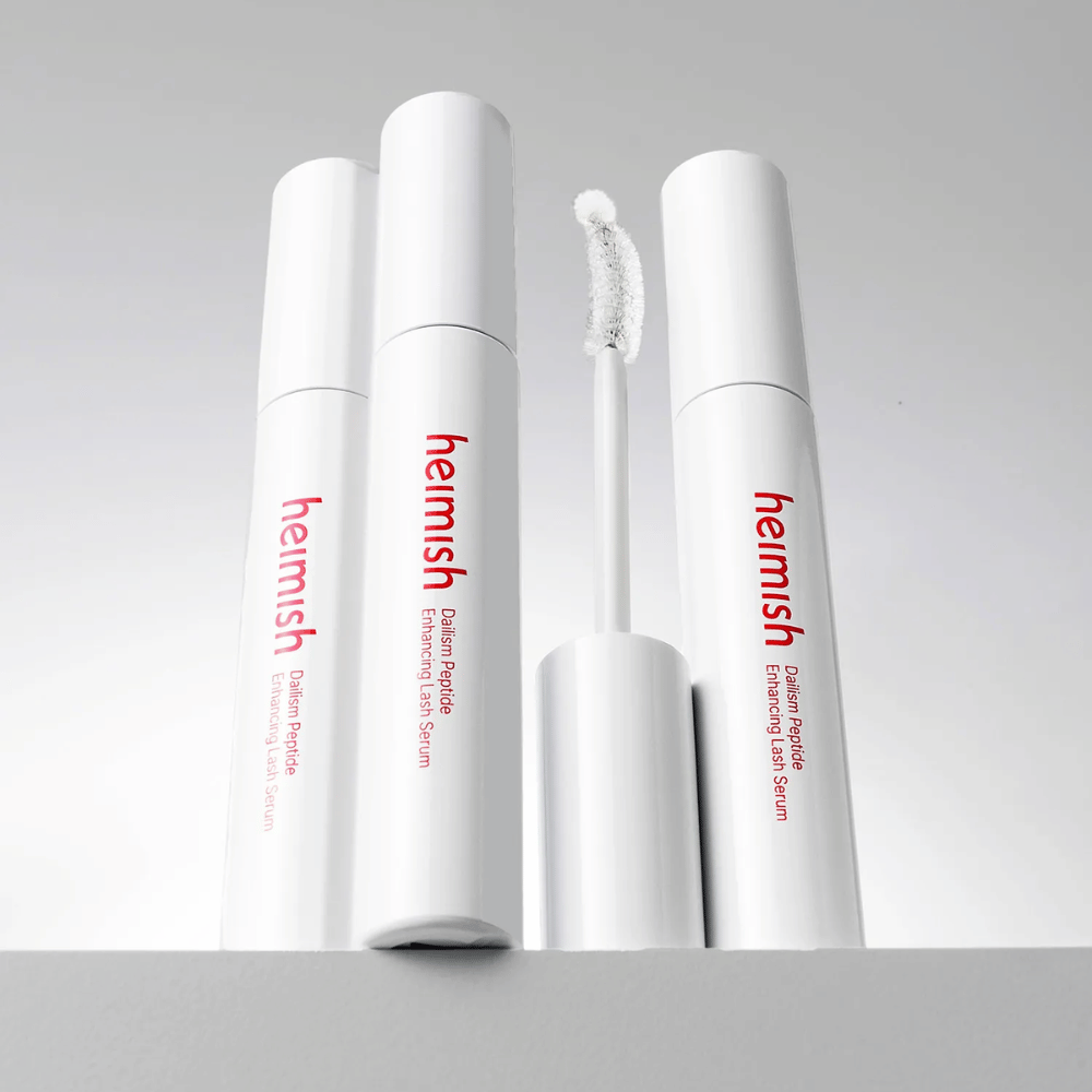 Várias embalagens de Heimish Dailism Peptide Enhancing Lash Serum são apresentadas juntamente com a escova aplicadora curva. A imagem destaca o design elegante do produto e a escova de precisão para uma aplicação fácil.