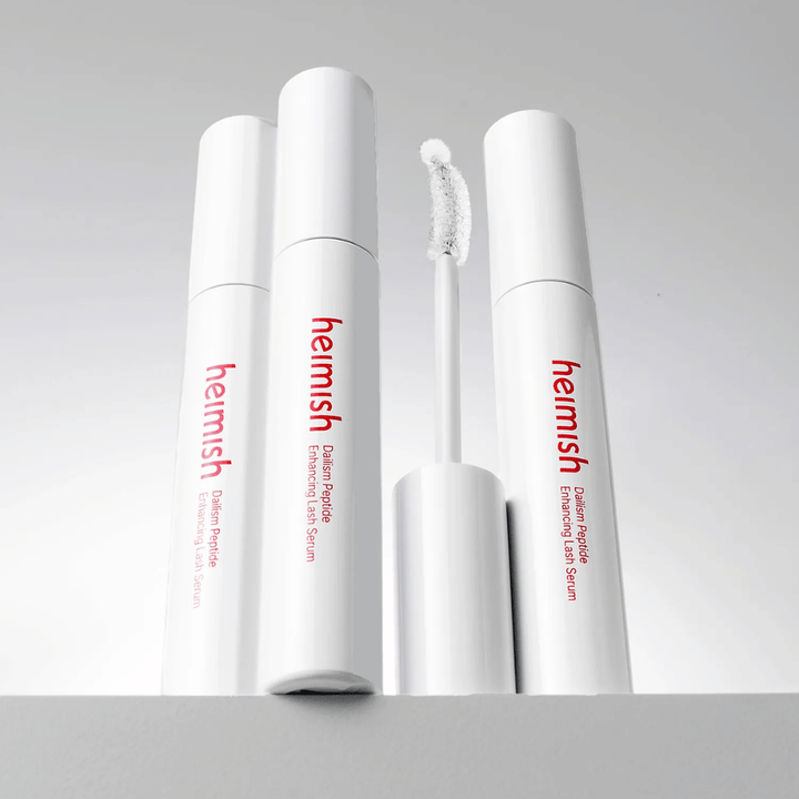 Várias embalagens de Heimish Dailism Peptide Enhancing Lash Serum são apresentadas juntamente com a escova aplicadora curva. A imagem destaca o design elegante do produto e a escova de precisão para uma aplicação fácil.