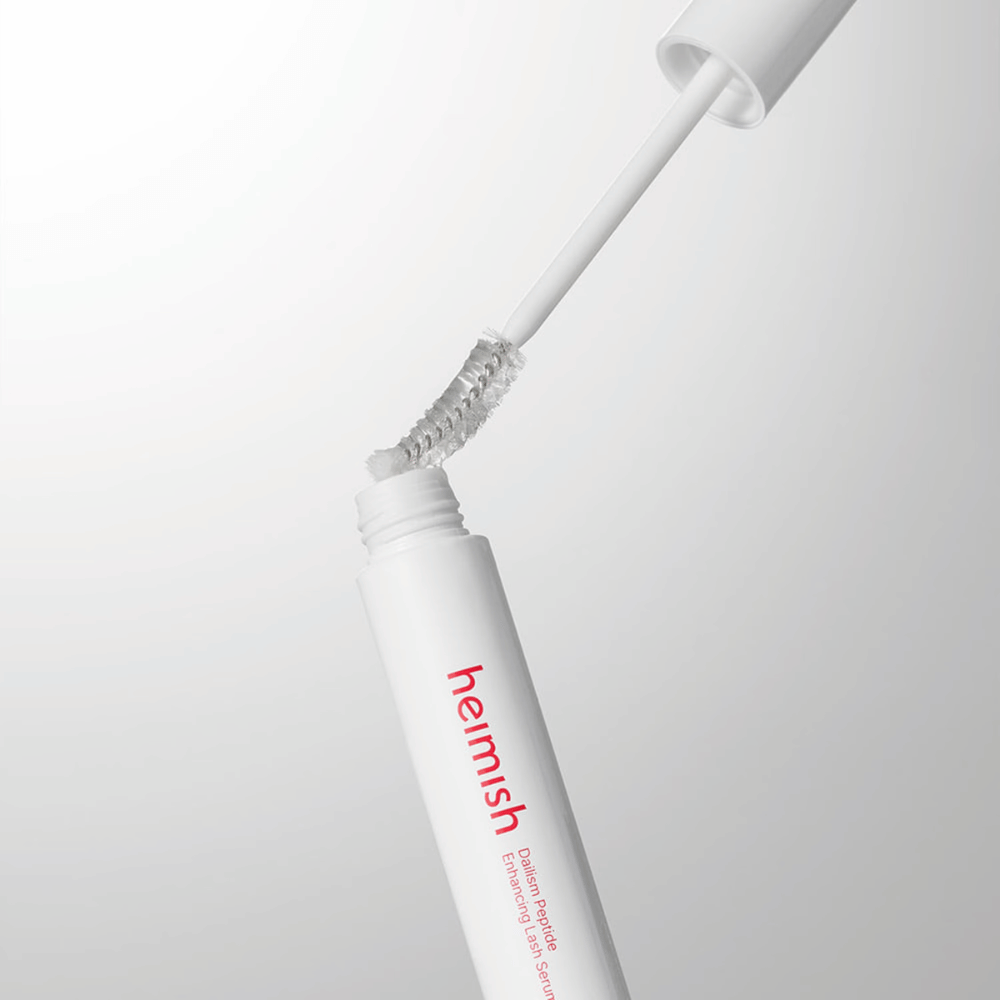 Grande plano do aplicador de Heimish Dailism Peptide Enhancing Lash Serum. A escova é retirada do recipiente branco e mostra a fórmula transparente do sérum que é aplicada nas pestanas.