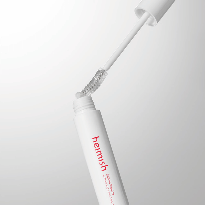 Grande plano do aplicador de Heimish Dailism Peptide Enhancing Lash Serum. A escova é retirada do recipiente branco e mostra a fórmula transparente do sérum que é aplicada nas pestanas.
