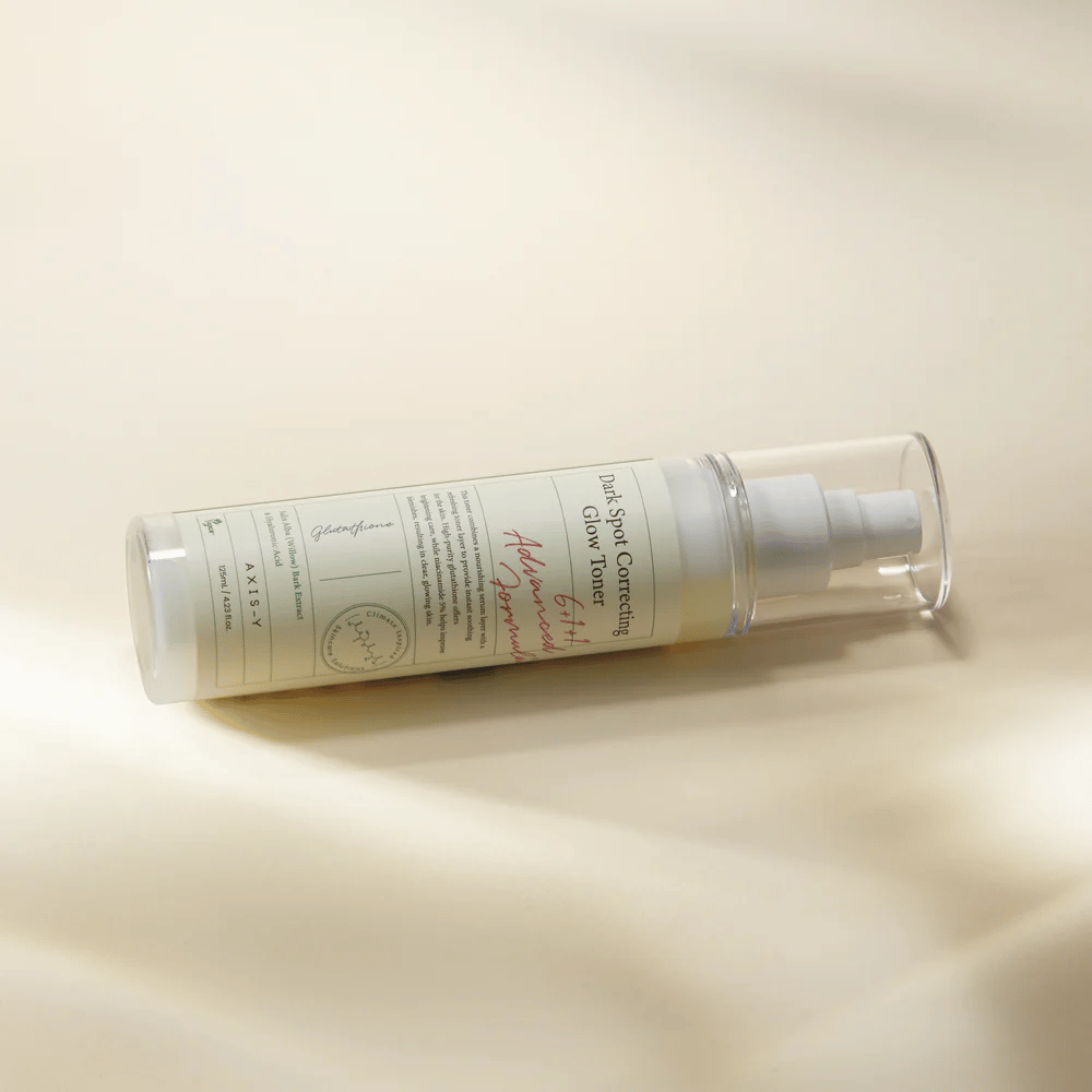 Uma garrafa deitada de "AXIS-Y Dark Spot Correcting Glow Toner" colocada sobre uma superfície clara. A garrafa é transparente com um rótulo branco e tem uma bomba de spray branca. O rótulo mostra informações sobre as propriedades iluminadoras e hidratantes do produto e contém ingredientes-chave como Glutationa e Niacinamida.