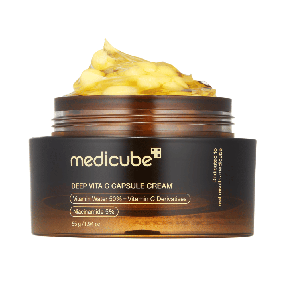 Uma imagem aproximada de "Medicube Deep Vita C Capsule Cream", mostrando a consistência do creme e as cápsulas de vitamina C. A embalagem é de cor marrom escuro com texto dourado e descreve o conteúdo de água vitaminada (50%) e niacinamida (5%), proporcionando uma apresentação visual das propriedades hidratantes e melhoradoras da pele do produto.