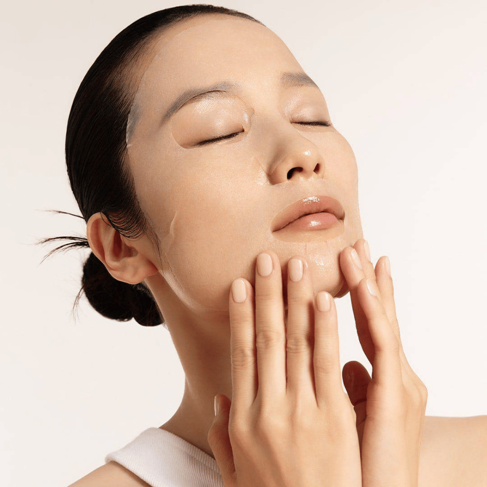 Modelo com pele hidratada e radiante aplica Sheet mask, foco no brilho, tom de pele uniforme e glow.