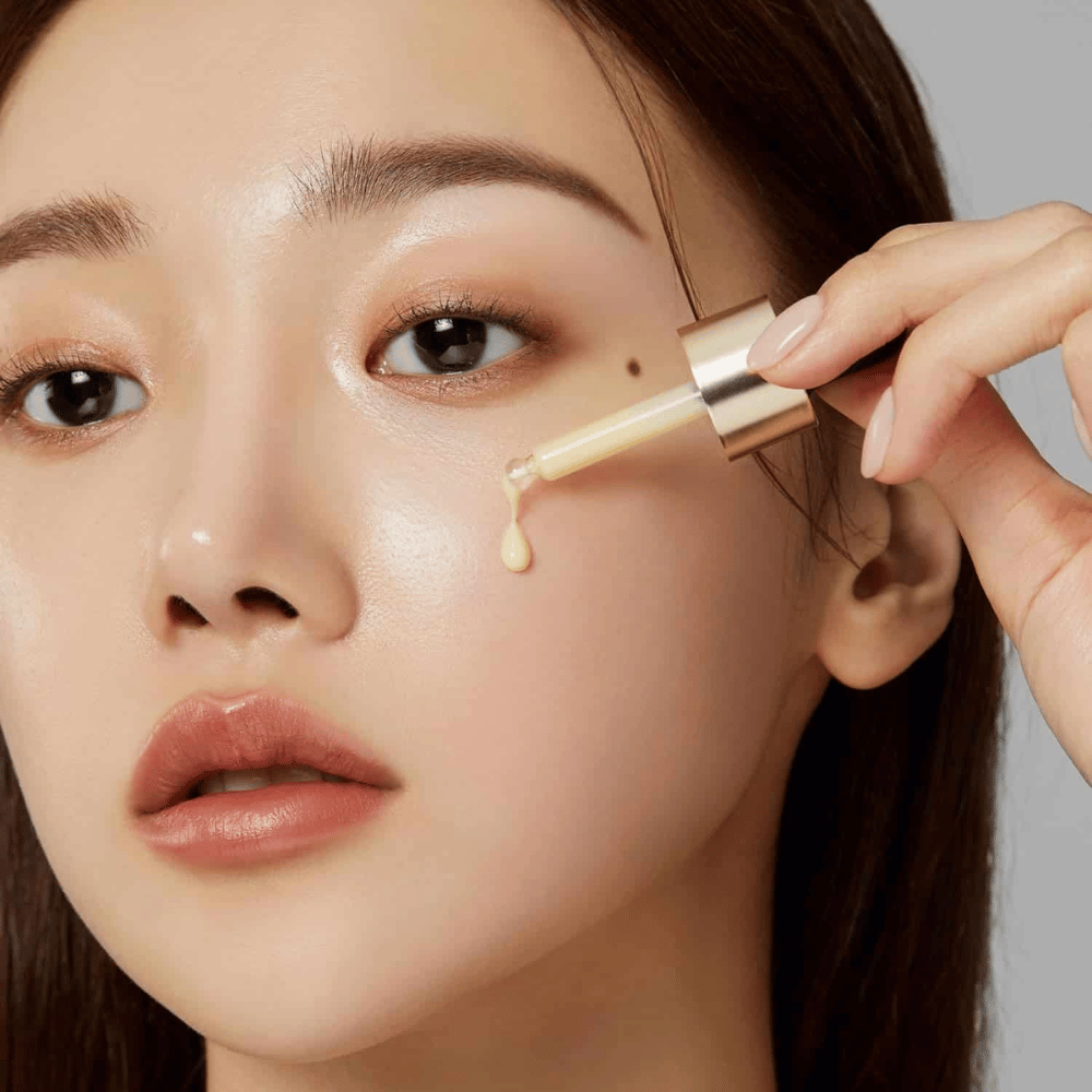 Close-up de uma mulher aplicando Medicube Deep Vita C Ampoule com um conta-gotas dourado. Uma gota do sérum está prestes a atingir sua pele, que tem um brilho natural. Perfeito para proporcionar um tom de pele mais claro e uniforme.