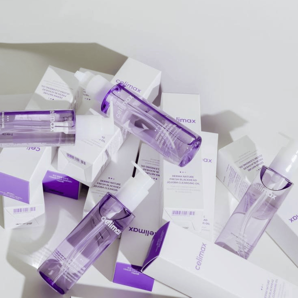 Celimax Derma Nature Fresh Blackhead Jojoba Cleansing Oil em frascos roxos colocados entre embalagens de produtos brancas com detalhes roxos.