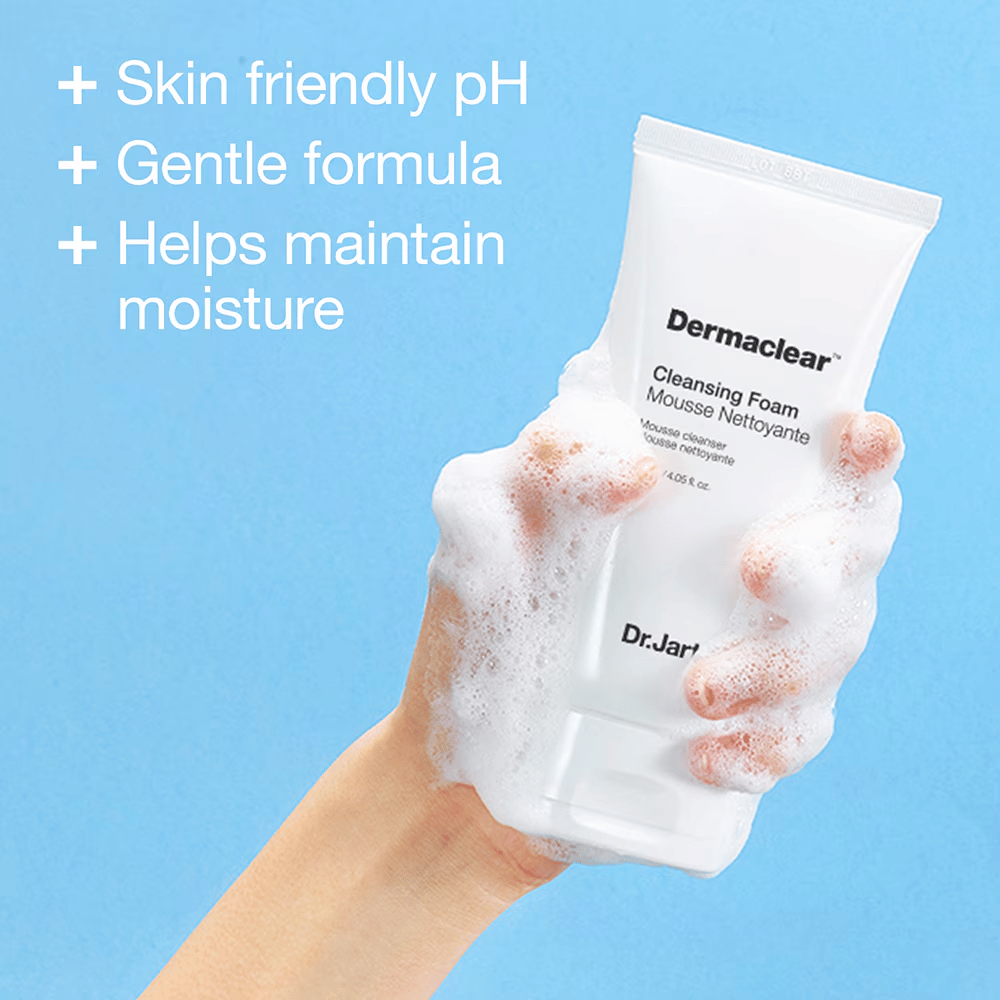 Mão coberta de espuma segurando Dr.Jart+ Dermaclear Cleansing Foam contra um fundo azul, com texto destacando pH amigável para a pele, fórmula suave e propriedades de retenção de umidade.