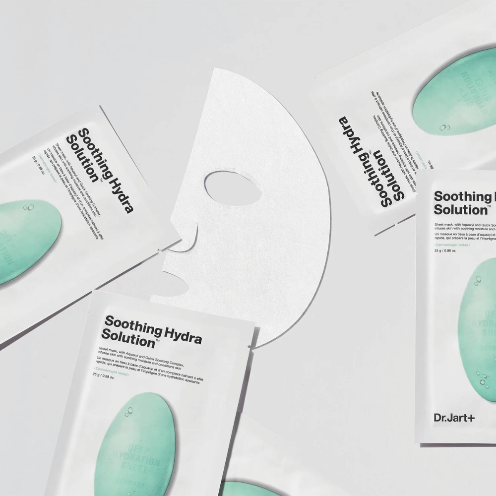 Várias embalagens de máscaras faciais "Soothing Hydra Solution" da Dr.Jart+ estão espalhadas sobre uma superfície clara. Uma máscara aberta está colocada no centro, mostrando sua estrutura branca e perfurada.