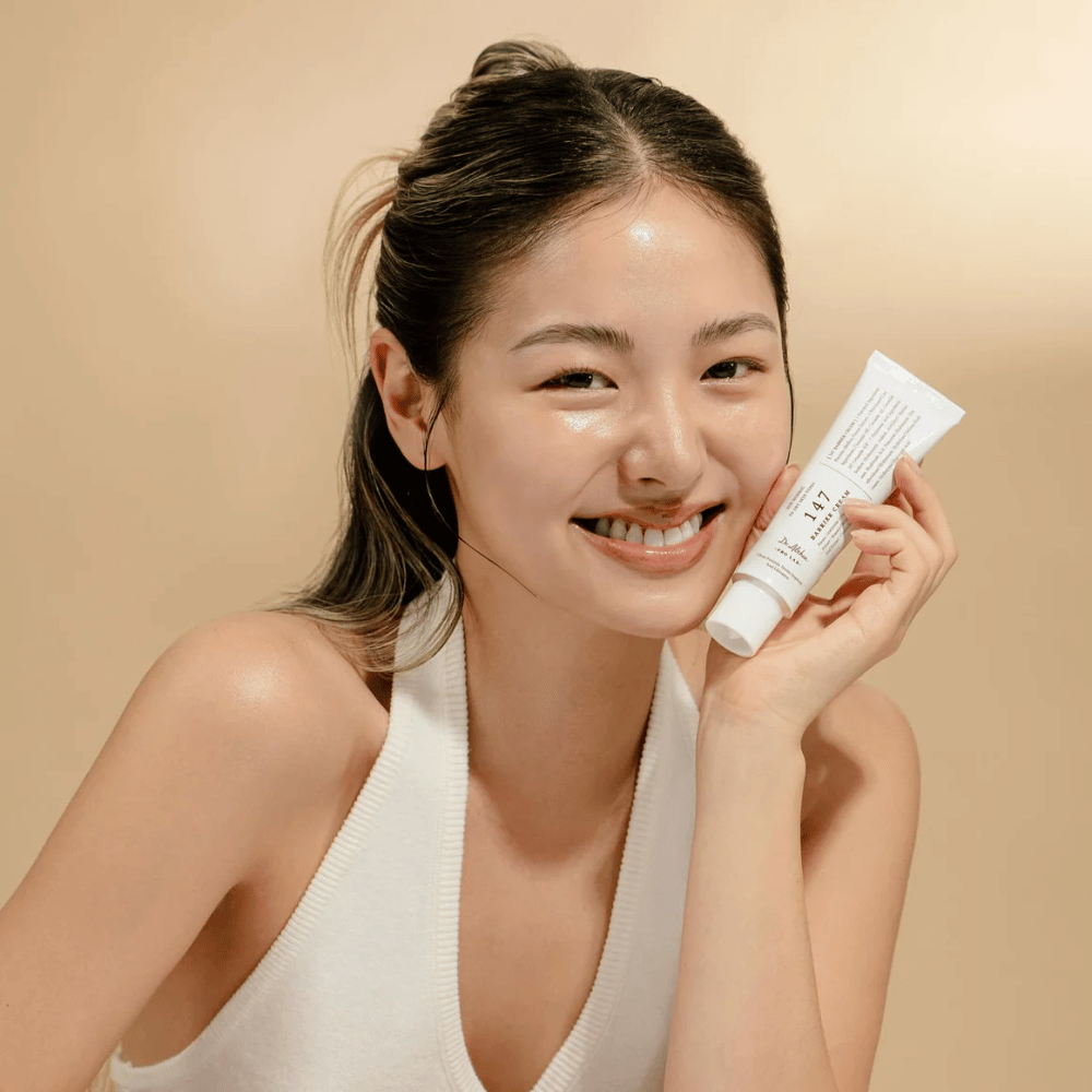 Modelo com pele luminosa sorrindo e segurando Dr. Althea 147 Barrier Cream – um creme facial coreano para barreira da pele e hidratação.