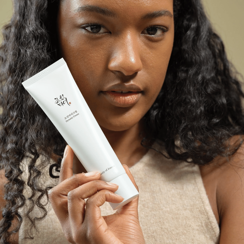 Uma mulher com cabelo encaracolado segura um tubo "Beauty of Joseon Dynasty Cream" perto do rosto, destacando o produto. O creme, apresentado em um elegante tubo branco, é projetado para nutrir e rejuvenescer a pele, enfatizando sua atratividade para diferentes tipos de pele.