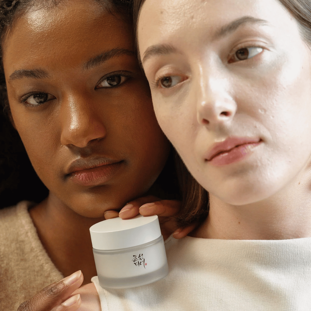 Duas mulheres, uma com tom de pele mais escuro e outra com tom de pele mais claro, estão próximas uma da outra com foco em seus rostos e um creme de pele da "Beauty of Joseon". O creme está colocado à frente delas, destacando seu uso para diferentes tipos de pele e sua capacidade de proporcionar brilho e nutrição.